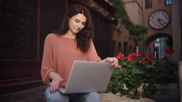 Beautiful happy cute joyful smiling romantic woman blogger working online to a laptop while studying outdoors, freelancer working while sitting on the decorative stone of a city street