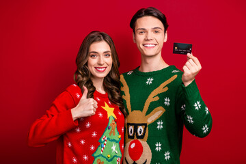 Photo of charming positive brother sister wear xmas sweaters holding credit card thumb up isolated red color background - obrazy, fototapety, plakaty