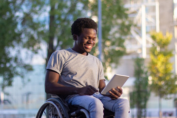 Young guy in wheelchair using his digital tablet in park. Happy Black man with disability enjoying video call on mobile device while spending time outdoors. Modern technology, disability concept. © KAMPUS