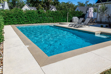 A rectangular new swimming pool with tan concrete edges in the fenced backyard of a new...