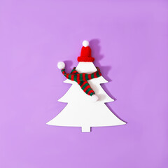A white Christmas tree with red and green striped winter scarf and a red hat, on lilac background. Minimal surreal concept for season greetings card or banner. Design for winter ski resort ad