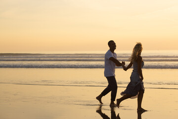 Fototapeta premium Affectionate couple walking on beach at sunset. Man and woman in casual clothes running along water at dusk. Love, family, nature concept