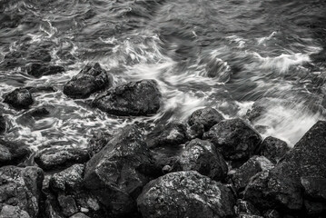 A high-angle, black and white long exposure shot of the sea. Water blurs as it washes over a rocky shoreline, creating a moody and dynamic texture with a monochromatic tone.
