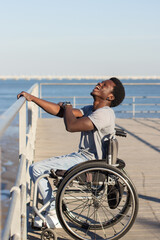 Fototapeta premium Cheerful Black man sitting in wheelchair at seafront on sunny day. Side view of confident guy with disability holding railing with his hand, and laughing happily. Disability, attitude concept.