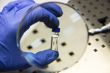 The hand of an expert in a rubber glove holds a vial under a magnifying glass