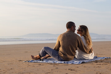 Tender couple at picnic at sunset. Back view of man and woman in casual clothes sitting on blanket in rays of setting sun. Picnic, meal, love concept