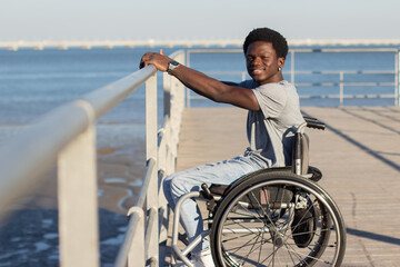 Happy Black man in wheelchair enjoying walking at seafront. Side view of confident smiling guy with paraplegia laying his hand on railing, smiling at camera. Full shot. Disability, attitude concept.