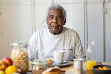 Portrait of senior man enjoying morning tea in kitchen. Serious grey-haired man calmly sitting at table posing for camera with joy when daughter preparing breakfast. Taking care of old people concept