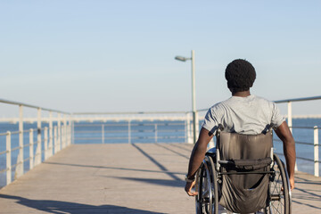 Young Black man riding wheelchair at seafront. Back view shot of man with physical disability...