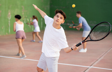 Athletic young hispanic guy playing frontenis on open court on summer day, hitting ball with strung tennis racquet to score to opposing team. Popular sports