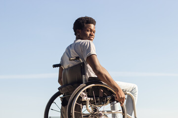 Black man in wheelchair enjoying sunny day in park, looking back and smiling at camera over the shoulder. Young African American guy with disability having fun outdoors. Disability, motivation concept