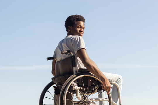 Black Man In Wheelchair Enjoying Sunny Day In Park, Looking Back And Smiling At Camera Over The Shoulder. Young African American Guy With Disability Having Fun Outdoors. Disability, Motivation Concept