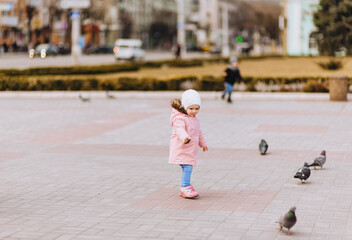 Fototapeta premium A little girl, a child in a pink coat, walks along the street in the city, feeding pigeons.