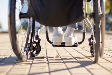 Person of unrecognizable gender and age in wheelchair spending time outside. Closeup back view shot of feet in white sneakers and wheels. Disability concept. - obrazy, fototapety, plakaty