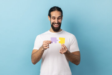 Portrait of man with beard in T-shirt holding two puzzle parts and smiling joyfully, ready to...