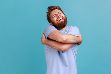 Portrait of satisfied positive handsome bearded man embracing himself, closed eyes and smiling with...