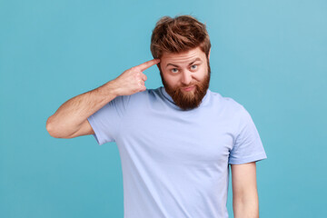 Fototapeta na wymiar Portrait of young bearded man showing stupid gesture with finger near head, out of mind, accusing crazy dumb plan, reckless expression. Indoor studio shot isolated on blue background.