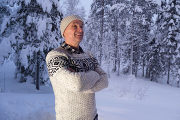 mature charismatic man 60 years in hat and Scandinavian sweater, smiling happily in winter forest against background of snowdrifts, concept of active lifestyle, good mood, enjoying winter nature