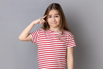 Are you kidding me? Portrait of little girl wearing striped T-shirt holding finger near head temple...