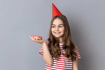 Portrait of happy joyful little girl wearing striped T-shirt and party cone, holding on palm tasty sweet donut, looking smiling at dessert. Indoor studio shot isolated on gray background.