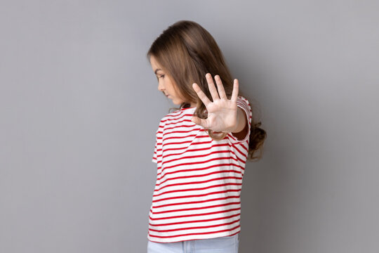 Portrait of scared serious little dark haired girl wearing striped T-shirt showing block stop gesture, set back bullying and violence. Indoor studio shot isolated on gray background.