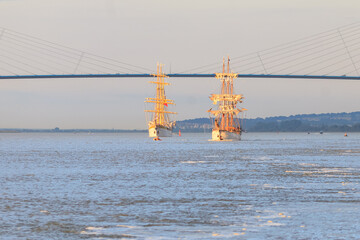 HONFLEUR, NORMANDY, FRANCE: Armada 2019 Grande Parade, tall ships Mircea and Le Français sailing in the Seine Estuary, in front of Normandy Bridge, with warm evening light - obrazy, fototapety, plakaty