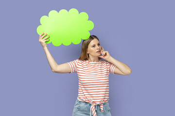 Portrait of pensive thoughtful young adult blond woman wearing striped T-shirt green bubble for text, looking away and holding her chin. Indoor studio shot isolated on purple background.