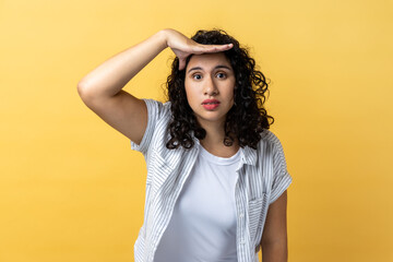 Portrait of attractive young adult woman with dark wavy hair keeps hand near forehead looks far away searches something on horizon. Indoor studio shot isolated on yellow background.