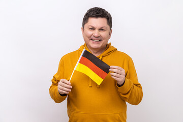 Satisfied positive middle aged man holding German flag in her hand and looking at camera with toothy smile, wearing urban style hoodie. Indoor studio shot isolated on white background.