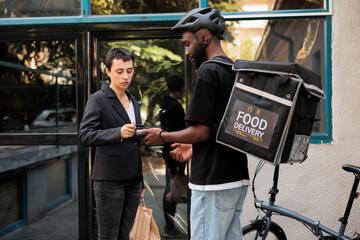 Office food delivery service, credit card pos terminal payment. African american courier on bike delivering restaurant takeaway meal, company employee paying contactless for lunch
