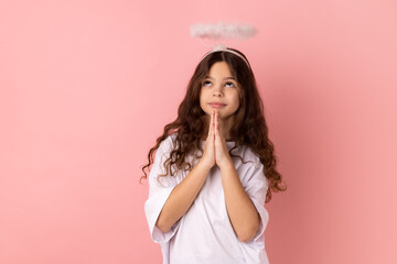 Portrait of little girl wearing white T-shirt keeps palms pressed together, pleading angel, has...
