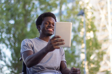 Fototapeta premium Black guy in wheelchair having video chat on tablet outside. Happy young man enjoying video call and smiling while holding mobile device and looking at screen. Disability, modern technology concept.