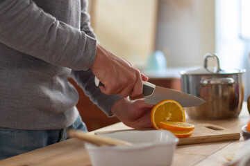 Side view of male hands slicing orange with knife. Unrecognizable man standing near table in home kitchen preparing fresh and healthy salad for dinner. Cooking food and healthy lifestyle concept