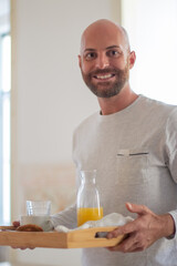 Happy homosexual man bringing his partner breakfast in bed. Vertical shot of smiling bearded man holding table tray with orange juice and buns. LGBT, food, morning routine concept