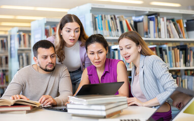Friendly group of students studying in the university library is preparing for classes on a laptop,...
