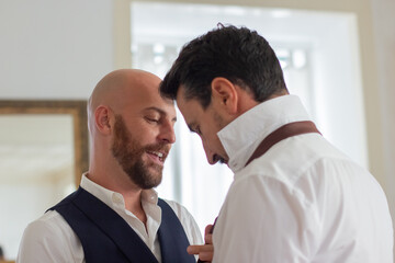 Gay tying his partner tie and talking with him in hotel room. Medium shot of handsome bearded man helping get dressed for wedding ceremony, spending time with lover. LGBT, love, marriage concept