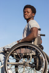 Confident Black man in wheelchair enjoying time outside. Happy African American young man in trendy clothes spending day outside in sunny weather. Vertical shot. Disability, attitude concept.