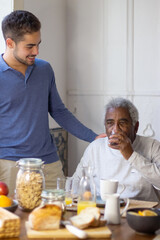 Portrait of happy granddad and grandson in cosy kitchen. Senior man drinking water and his smiling grandson hugging him and talking to him. Taking care and providing support to old people concept