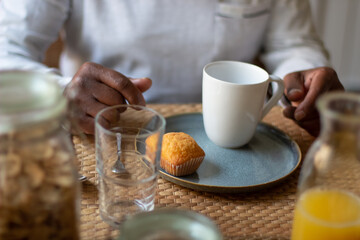Close-up of aged mans hands on table during having breakfast. Old Latin man sitting at table with cake and cup on it, going to drink tea prepared by daughter. Happy life, care for aged people concept