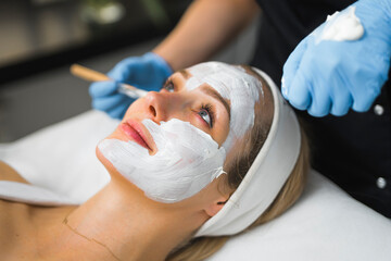 Indoor closeup portrait of the procedure of spreading deeply cleansing white facial mask onto the face of beautiful blond young adult caucasian woman by an unrecognizable specialist in protective