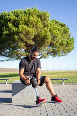 Young sportsman with disability sitting on stone bench. Thoughtful bearded man with artificial leg sitting outdoors attaching prothesis for training. Active life of people with disability concept