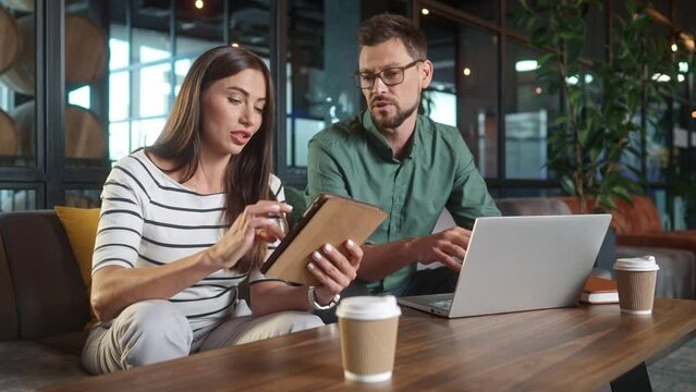 Happy successful beautiful woman using tablet working online and handsome busy man working on laptop in cafe. Concentrated cheerful male and female talking about job laughing. Freelance concept.