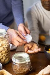 Close-up of young mans hands giving medicine to granddad. Senior Latin man sitting at table with food on it and grandson giving him daily medicine. Taking care, providing support to old people concept