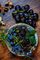 Top view berries blue blackberry grapes on a wooden table and on a small golden plate. Green leaves and dry flowers. Shallow depth of field. Dark blurred background. Copy space.