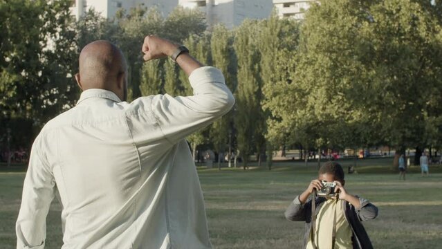 Black man showing biceps while his young son taking photo. African American boy using camera, taking picture of his strong father while spending time in park. Photography, parenthood, father concept.