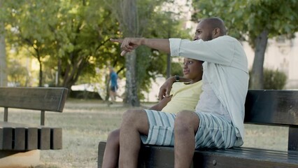 Father pointing at something while sitting on bench with his son on warm summer day. Black middle-aged man and young boy watching at something in distance while resting in park. Father and son concept
