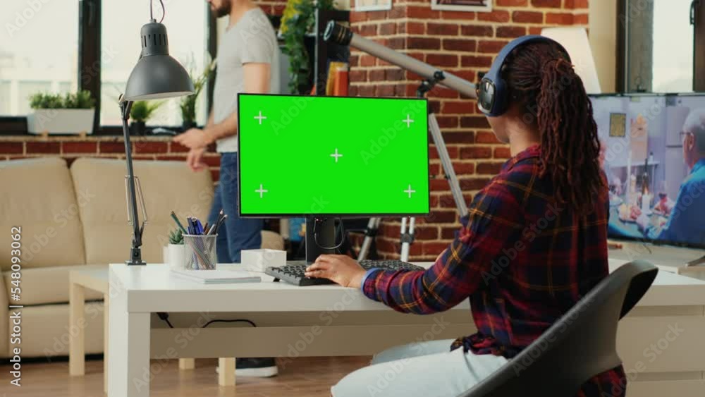 Poster office employee wearing headphones and using greenscreen on computer desktop, sitting at desk and wo