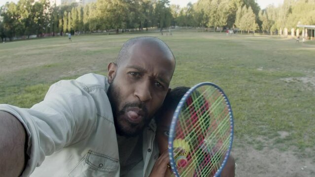 Father and son taking selfie in park, having fun together. Black man and young boy making funny faces. Child holding badminton shuttlecock and racket. Camera POV. Father and son, leisure concept.