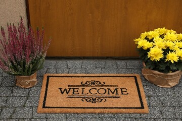 Doormat with word Welcome and beautiful plants on paving stone near entrance