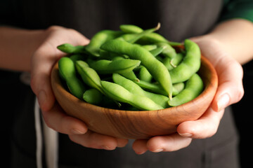Woman holding bowl with green edamame beans in pods on black background, closeup